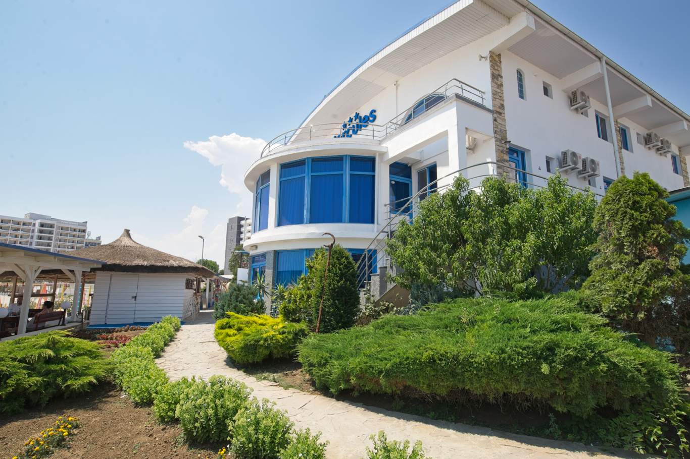 Vedere frontală a Hotelului Hercules cu fațadă albă și albastră, grădină îngrijită și intrare principală