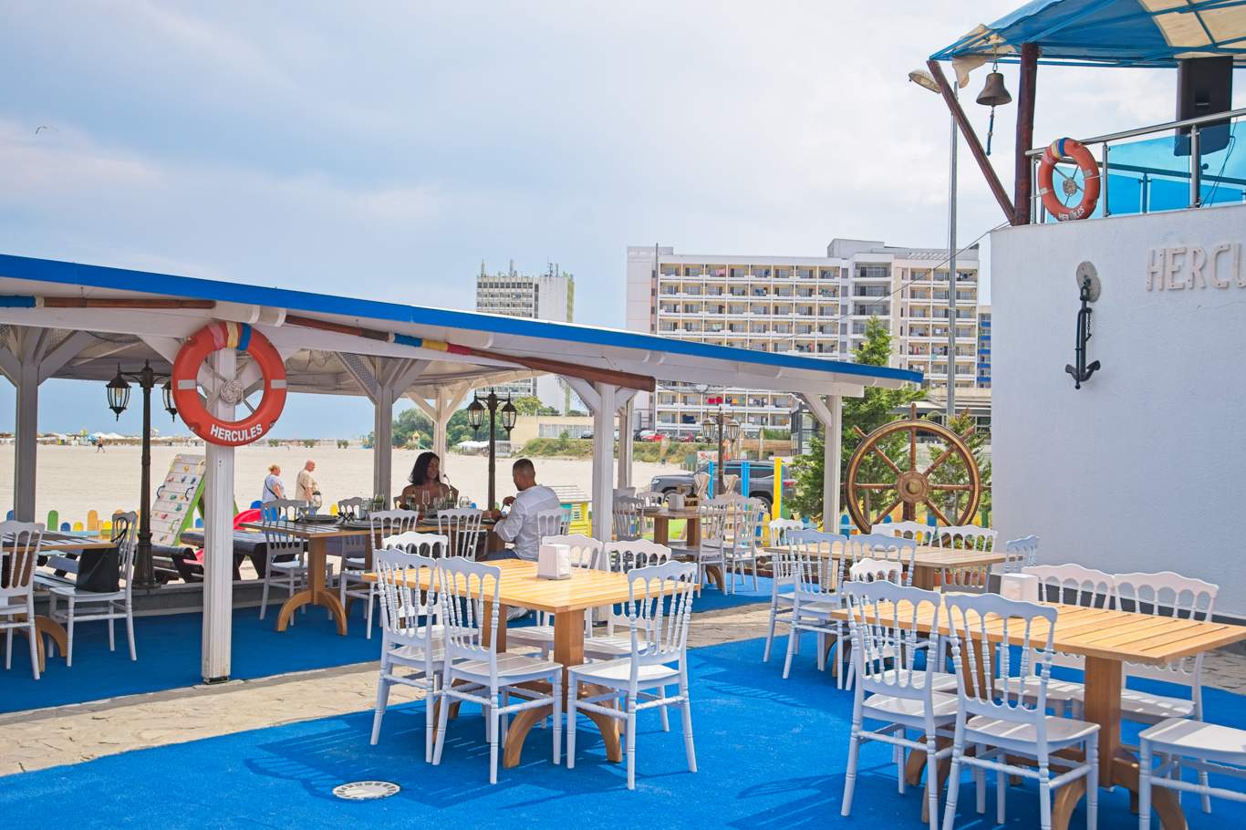 Terasă exterioară a restaurantului Hotel Hercules cu decor nautic, colac de salvare, cârmă de corabie și mese albe cu scaune, cu plaja în fundal