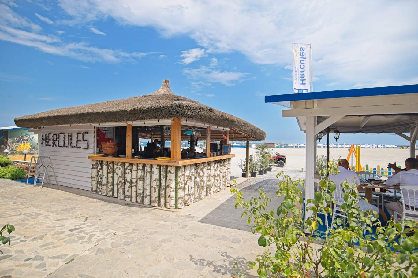Vedere frontală a barului de plajă Hotel Hercules cu acoperiș din stuf și nisip în față
