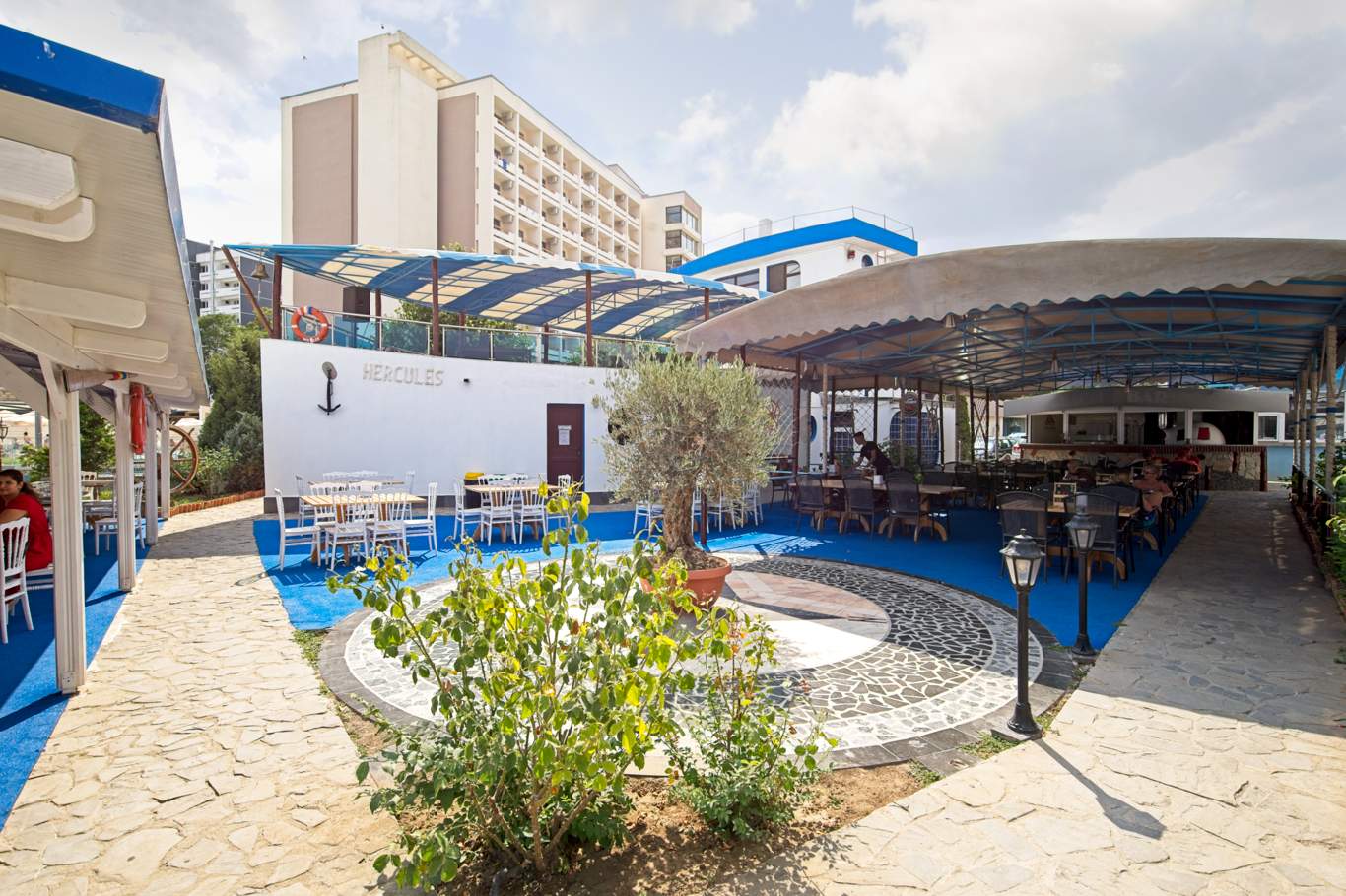 Vedere de ansamblu a terasei restaurantului Hotel Hercules cu mese și scaune, alee pietonală cu mozaic, vegetație ornamentală și sigla Hercules pe peretele alb