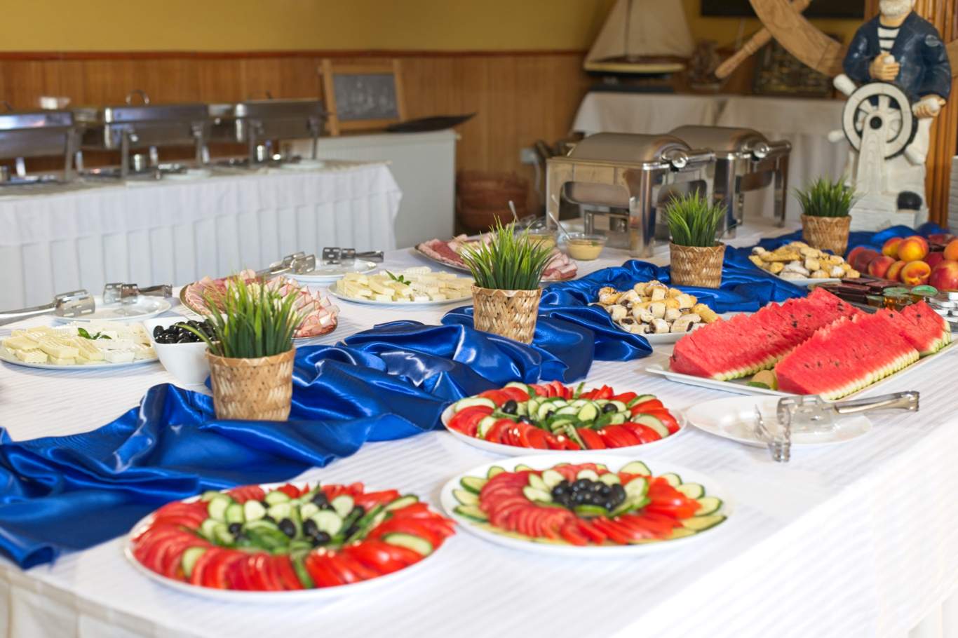 Masă de bufet bogat decorată cu platouri de legume, fructe și mezeluri, fețe de masă albastre și decor nautic în restaurantul Hotel Hercules