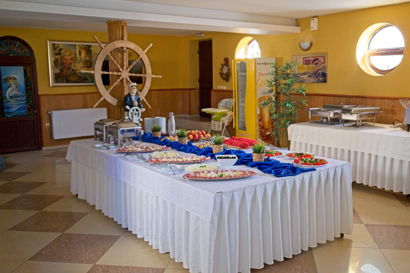 Masă bufet pentru micul dejun în restaurantul Hotel Hercules, cu preparate variate pe fețe de masă albastre, decor nautic cu cârmă de corabie și statuetă de marinar pe pereți galbeni