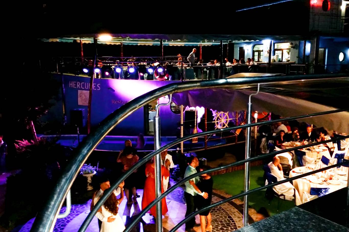 Vedere nocturnă a terasei restaurantului Hotel Hercules în timpul unui eveniment, cu lumini de scenă colorate, oaspeți la mese, pistă de dans și scenă cu muzicieni