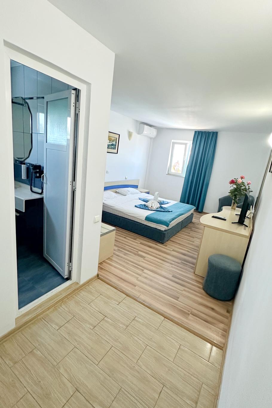 Cameră cu pat, perdele turcoaz, parchet din lemn și baie en-suite vizibilă