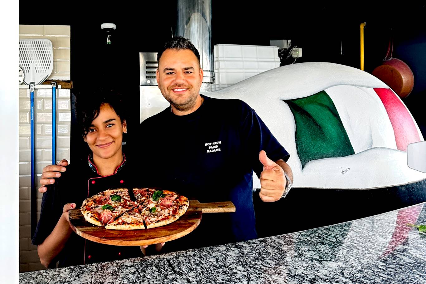 Membrii echipei Hotelului Hercules prezentând o pizza proaspătă pe blat de lemn, în fața cuptorului tradițional cu lemne