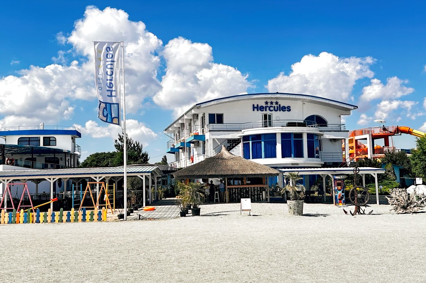 Vedere de ansamblu a complexului Hotel Hercules cu terase, umbrar și plajă, pe timp însorit